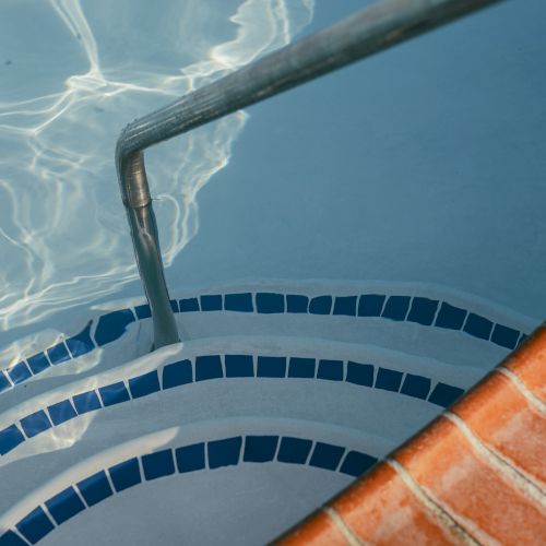 This image shows the steps of a swimming pool with a metal handrail, surrounded by brick tiles and water with sunlight reflections.