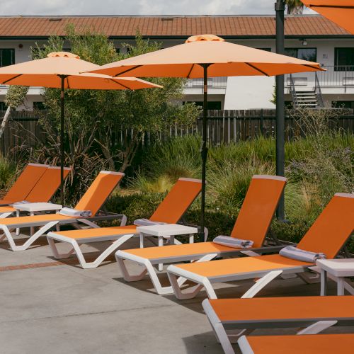 A row of orange lounge chairs with matching umbrellas by a poolside, surrounded by greenery and a building in the background.