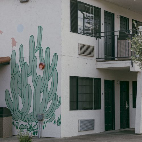 A two-story white building with a green cactus mural on the side. It has a red tile roof and black railings on the upper level walkway.