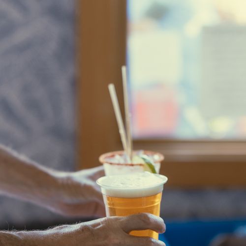 A person holds a plastic cup filled with a beverage, possibly beer, near a window; another cup with straws is seen in the background.