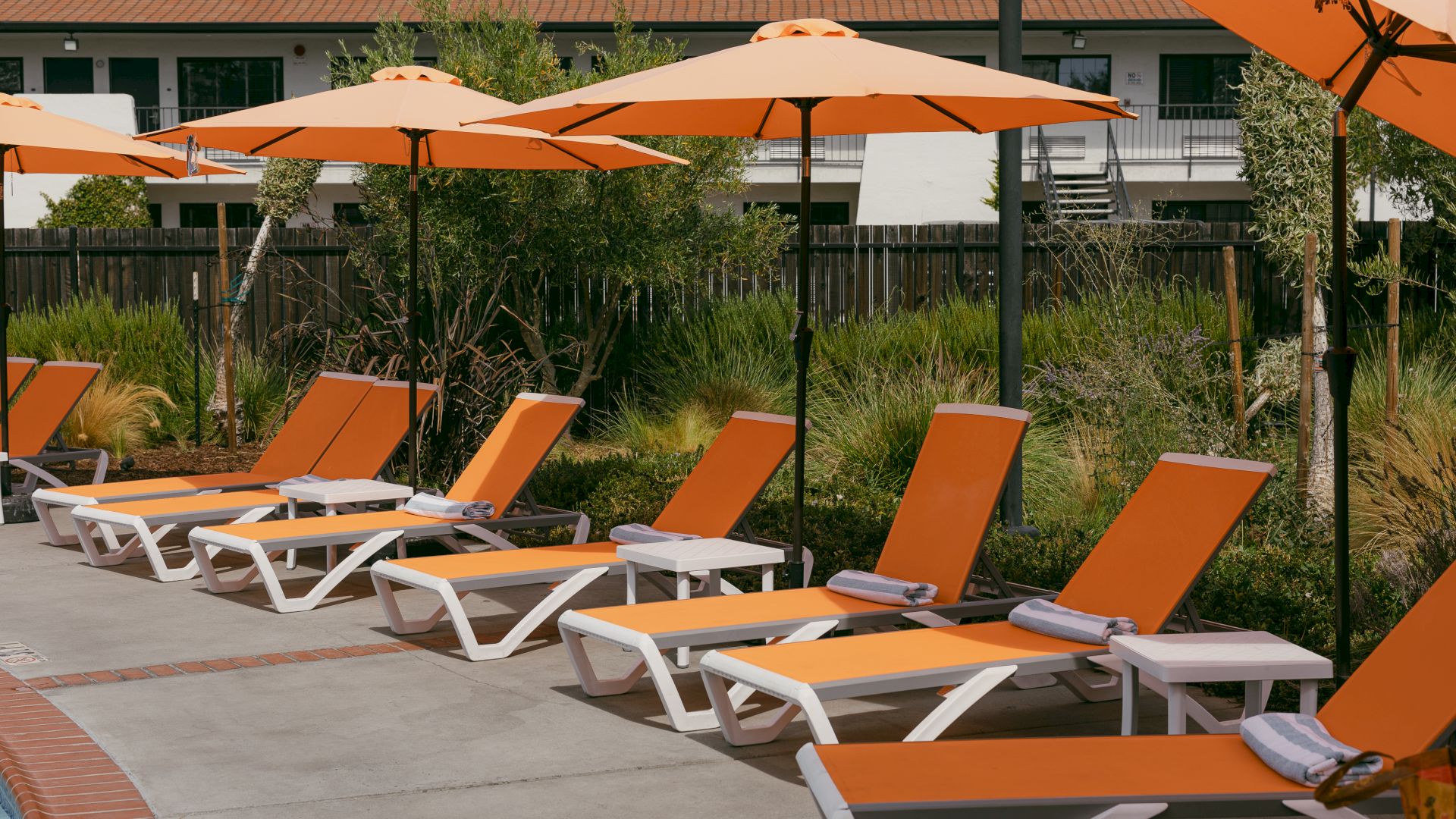 A row of orange lounge chairs and umbrellas beside a pool with a building in the background.