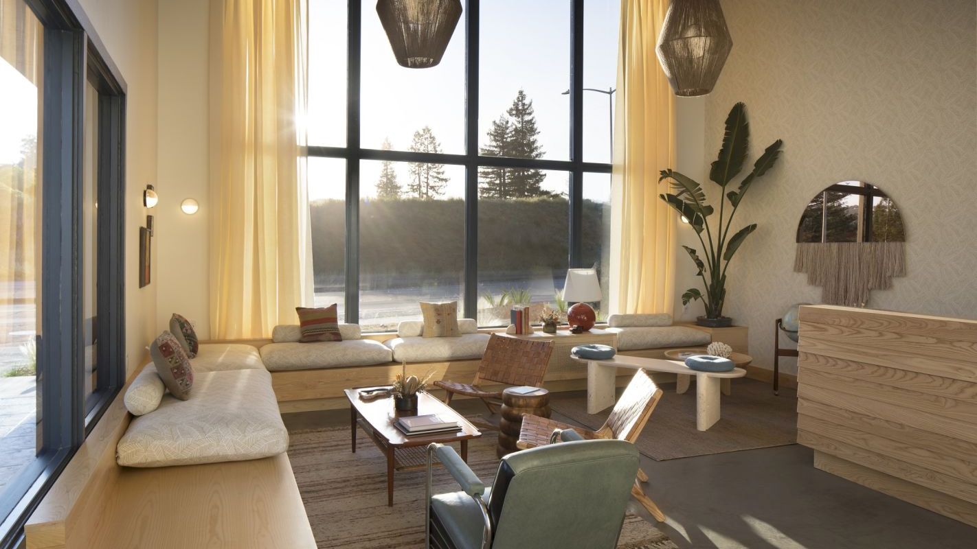 A cozy, well-lit living room with large windows, modern furniture, hanging pendant lights, and a large plant.