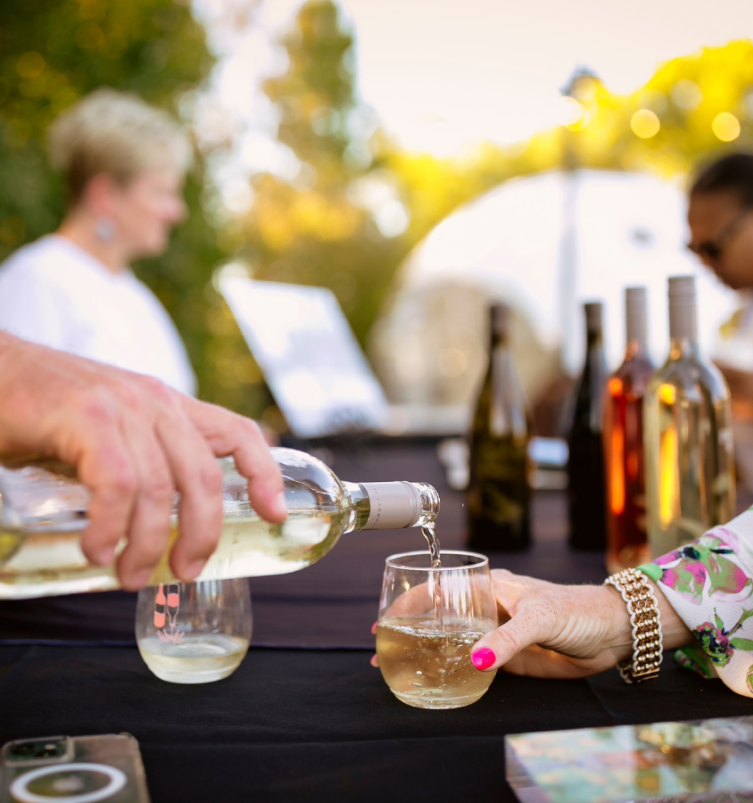 A person pours white wine into a glass for another hand, with bottles and people in the background and a phone on the table, ending the sentence.