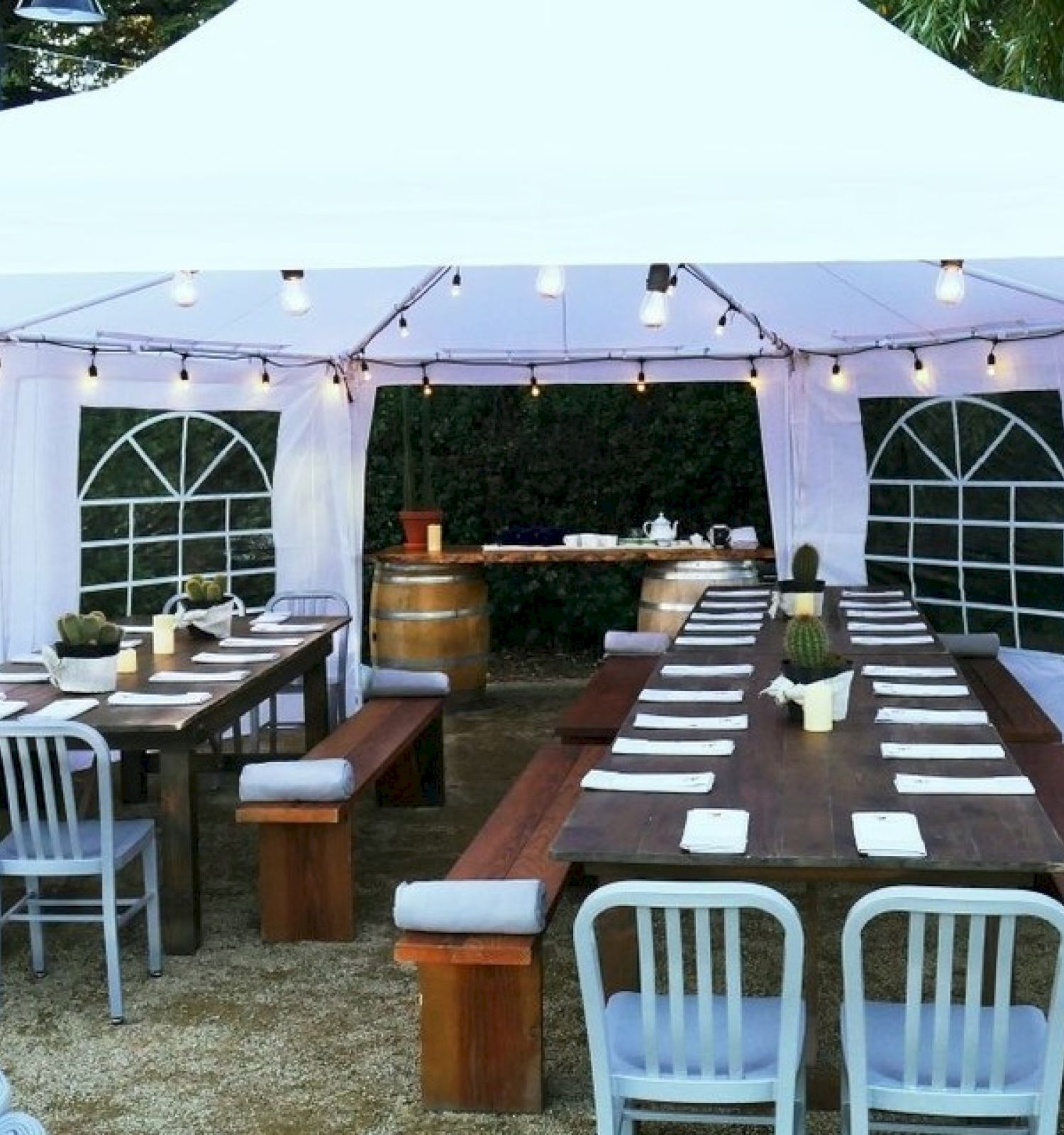 An outdoor dining setup with wooden tables, benches, and chairs under a white canopy, decorated with string lights, surrounded by greenery.