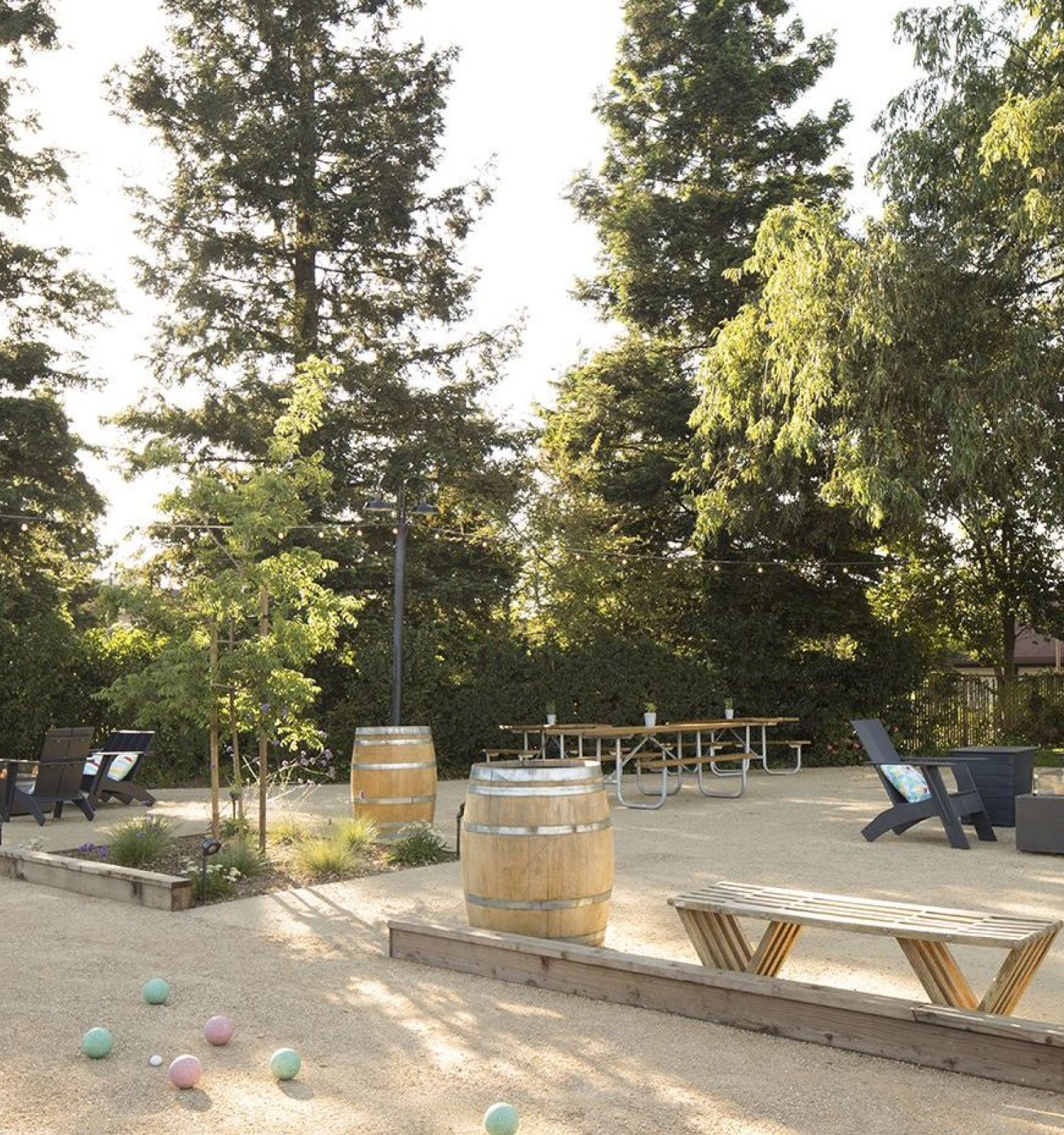 A serene outdoor area with lounge chairs, wooden barrels, a bench, a table for seating, and colorful balls, all surrounded by tall trees and greenery.