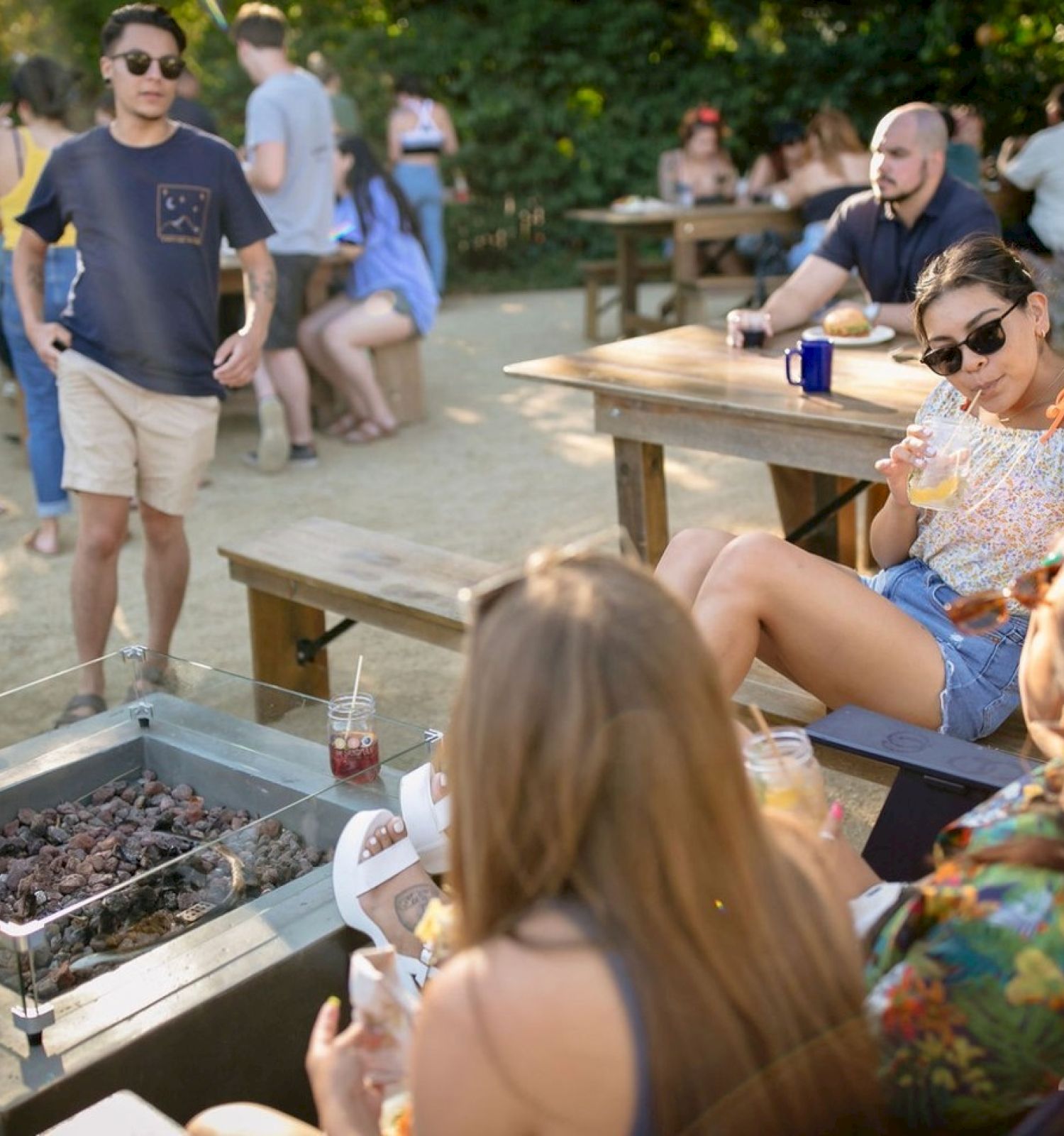 A group of people enjoying a casual outdoor gathering with drinks and snacks around a fire pit on a sunny day.