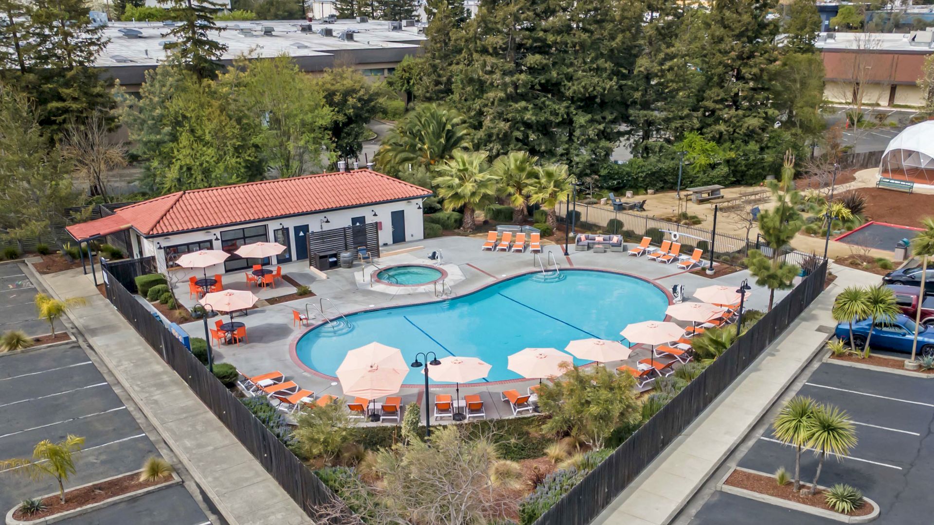 An aerial view of a swimming pool area with umbrellas and lounge chairs, surrounded by a parking lot and trees.