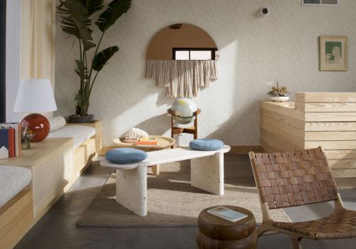 A cozy room with a wooden bench, two stools, a mirror with fringe, a globe, a lamp, books, and a woven chair on a rug, creating a warm atmosphere.