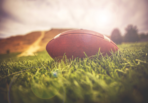 A football lies on a grassy field under a bright sky, with distant blurred trees and hills in the background.