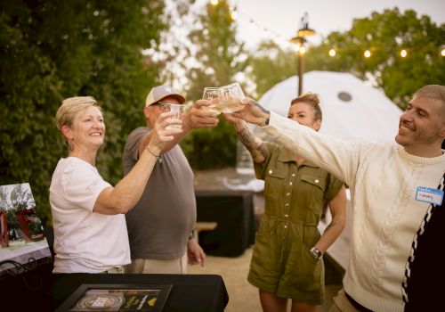 A group of four people outside, toasting and smiling with drinks in hand, with string lights and trees in the background.