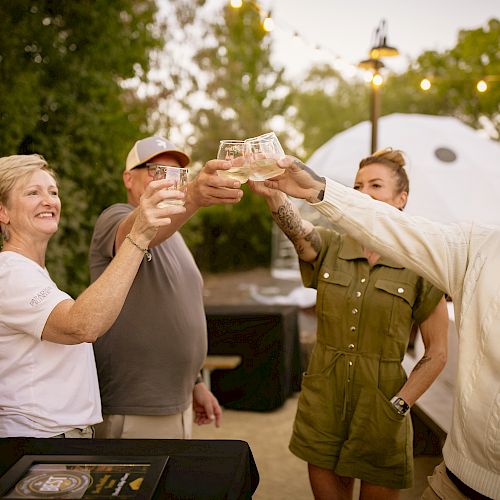 A group of four people outside, toasting and smiling with drinks in hand, with string lights and trees in the background.