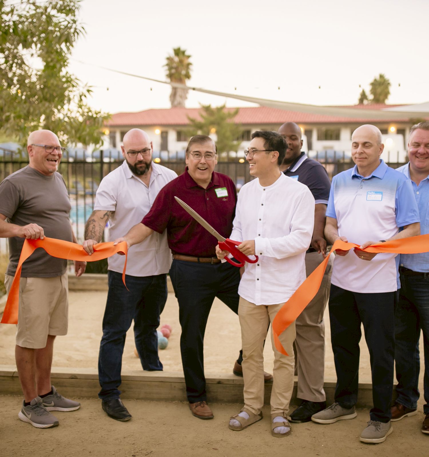 A group of people stands together outdoors, holding an orange ribbon and a pair of large scissors, presumably for a ribbon-cutting ceremony, ending the sentence.