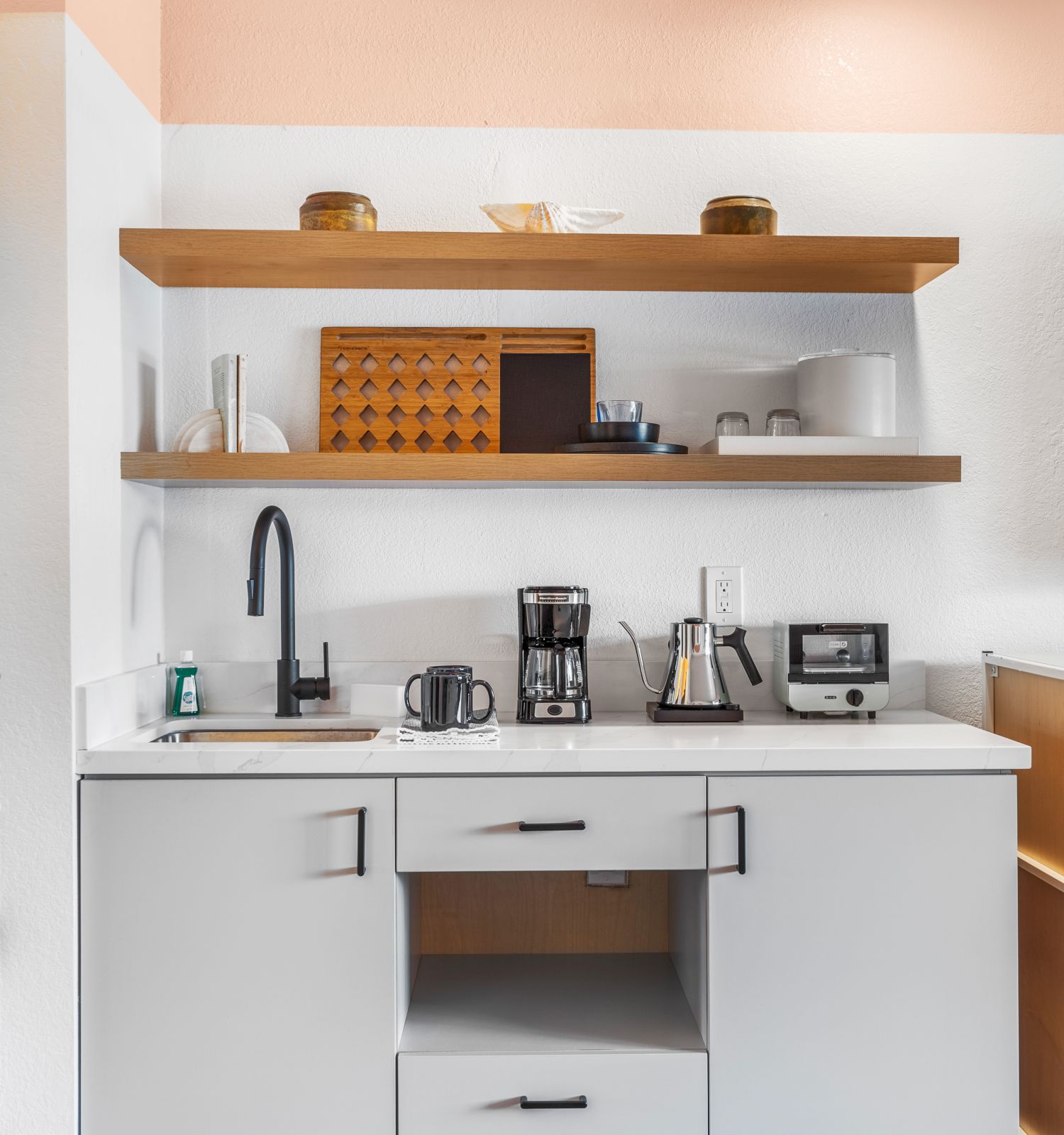 A modern kitchenette with open shelves, a sink, coffee maker, and other appliances. There's a flat-screen TV next to a mini-fridge.