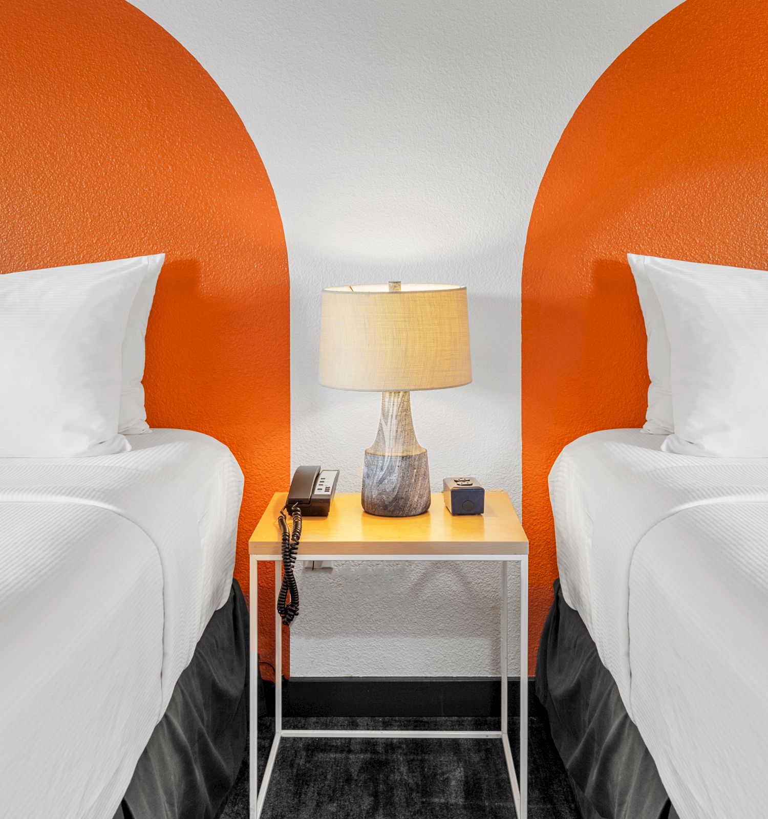 The image shows two white beds with pillows, separated by a small nightstand with a lamp, phone, and decor. Orange semicircles are painted on the wall.