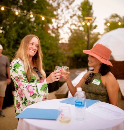 Two women toast at a table outdoors. Other people are in the background, and there are string lights above.