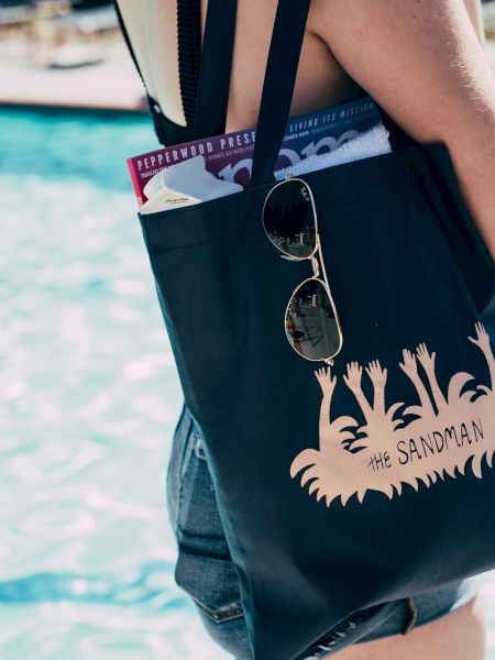 A person by the pool carries a black bag with designs, sunglasses clipped on, and magazines inside. They wear shorts and a sleeveless top.