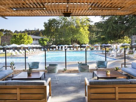 Outdoor seating area overlooking a swimming pool with shaded lounge chairs, tables, and plants, surrounded by trees and buildings in the background.