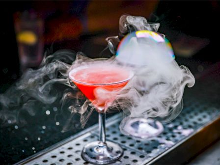 A red cocktail in a glass, emitting smoke or vapor, placed on a bar counter with a smoky, atmospheric effect in the background.