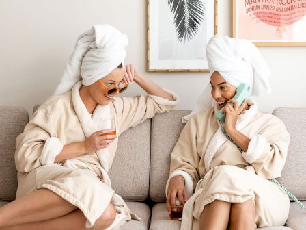 Two people are sitting on a couch wearing bathrobes and towels on their heads, one talking on a phone and the other holding a drink and wearing sunglasses.