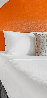 This image shows two beds with white linens and decorative pillows, separated by a small nightstand with a lamp and other items, against an orange backdrop.