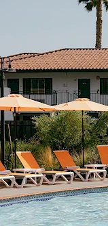 A poolside scene with orange lounge chairs and umbrellas, set against a building with balcony railings and a tiled roof in the background.