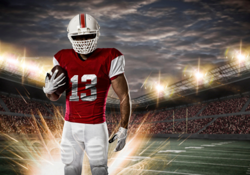 A football player in red and white gear holds a football on a lit stadium field with a cloudy sky and bright lights in the background.