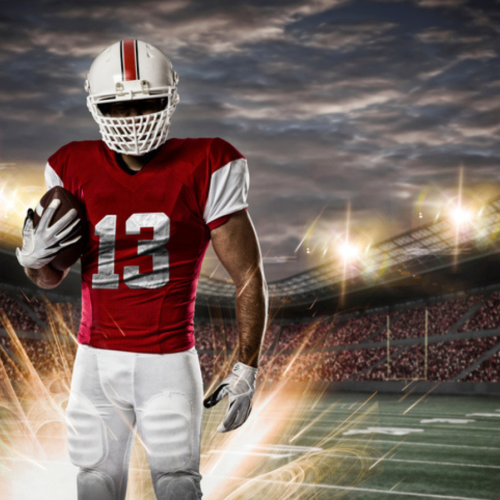 A football player in red and white gear holds a football on a lit stadium field with a cloudy sky and bright lights in the background.