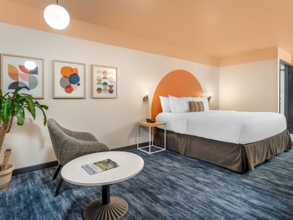 A contemporary hotel room with a bed, modern art, a chair, table, and plant, featuring warm tones and stylish decor.
