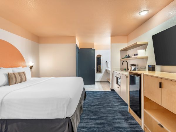 A modern hotel room with a large bed, nightstand, wall-mounted TV, kitchenette, and an open bathroom with a sink and mirror.
