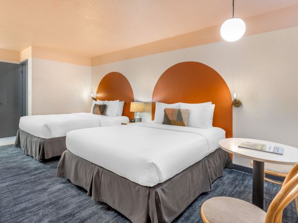 A hotel room with two beds, orange wall accents, a side table, chairs, and soft lighting.