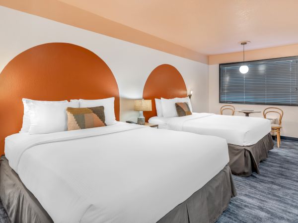 The image shows a hotel room with two beds, orange circular wall decor, a window, and a small table with chairs.