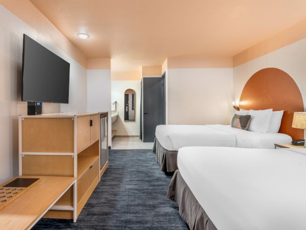 A hotel room with two beds, a wall-mounted TV, a wooden shelf, and a lamp on a nightstand is displayed in the image.