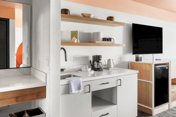 The image shows a modern kitchenette with open shelving, a coffee maker, a kettle, a TV on a stand, and a mini-fridge, all in a bright, organized space.