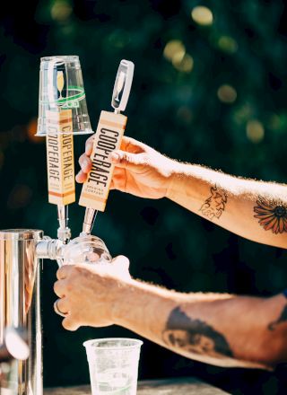 A person with tattoos is dispensing beer from taps labeled "GOOD VIBES" into a clear plastic cup outdoors.