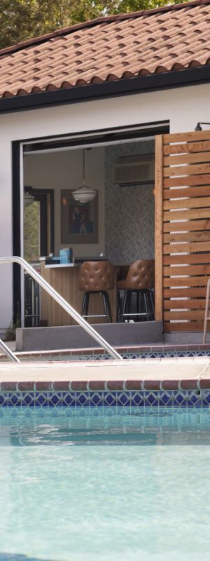 A serene outdoor pool area with blue umbrellas, loungers, railings, and a building with a tiled roof and open seating area.