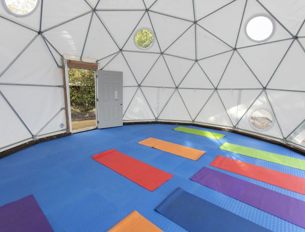 A geodesic dome structure with yoga mats laid out on a blue floor, an open door, and natural light coming in from circular windows, ending the sentence.