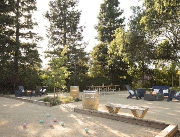 The image shows an outdoor area with trees, seating arrangements, and a few bocce balls scattered on the ground, suggesting a recreational space.