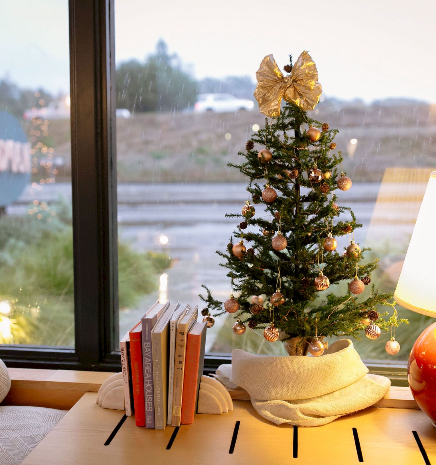 A small decorated Christmas tree sits by a window with books and a lamp on a cozy table, looking out onto a rainy scene.