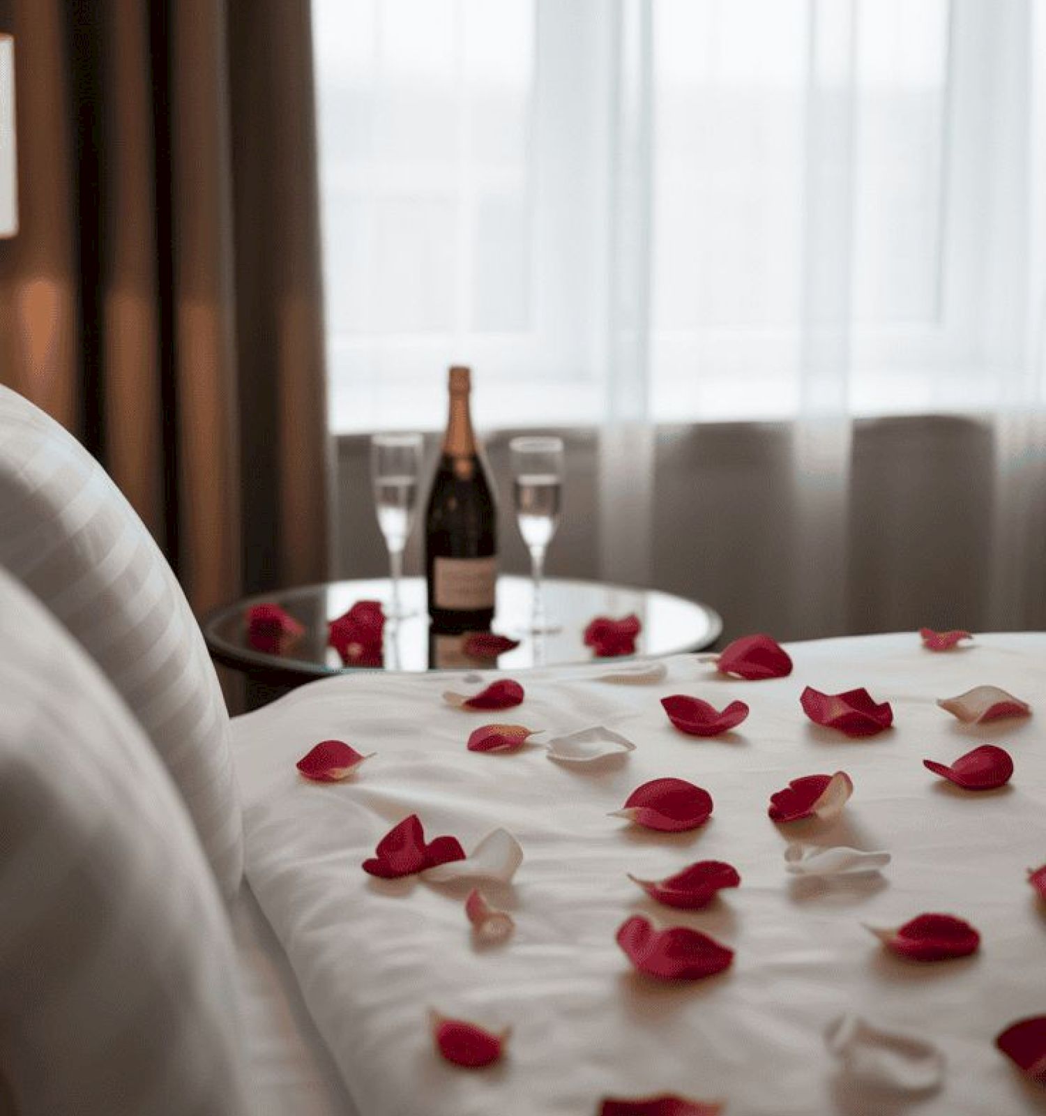Aromantic hotel room scene with a bed strewn in red rose petals, a champagne bottle and glasses on the table, soft natural light filtering through sheer curtains.