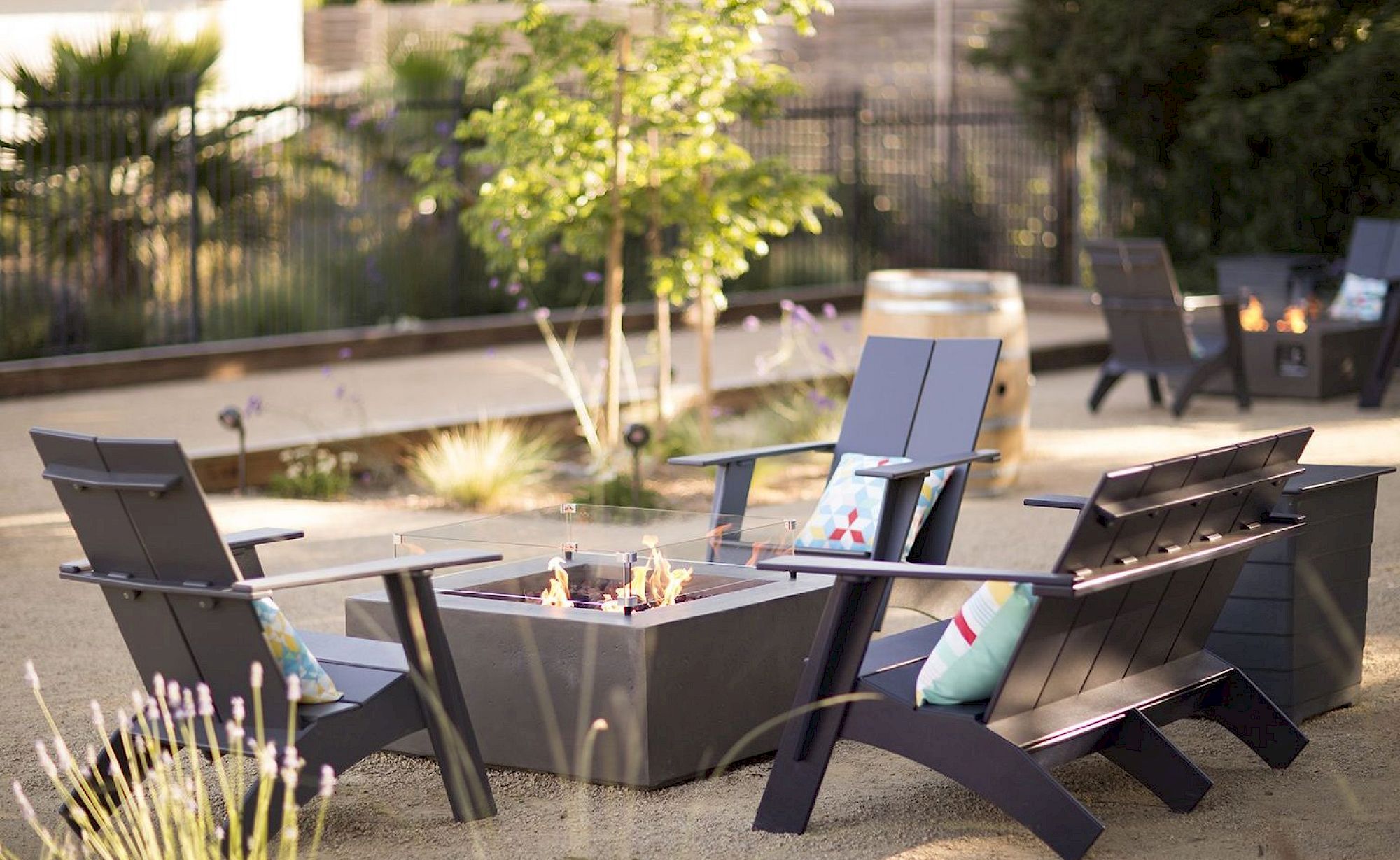 Several outdoor chairs surround a square fire pit in a landscaped garden, creating a cozy sitting area.