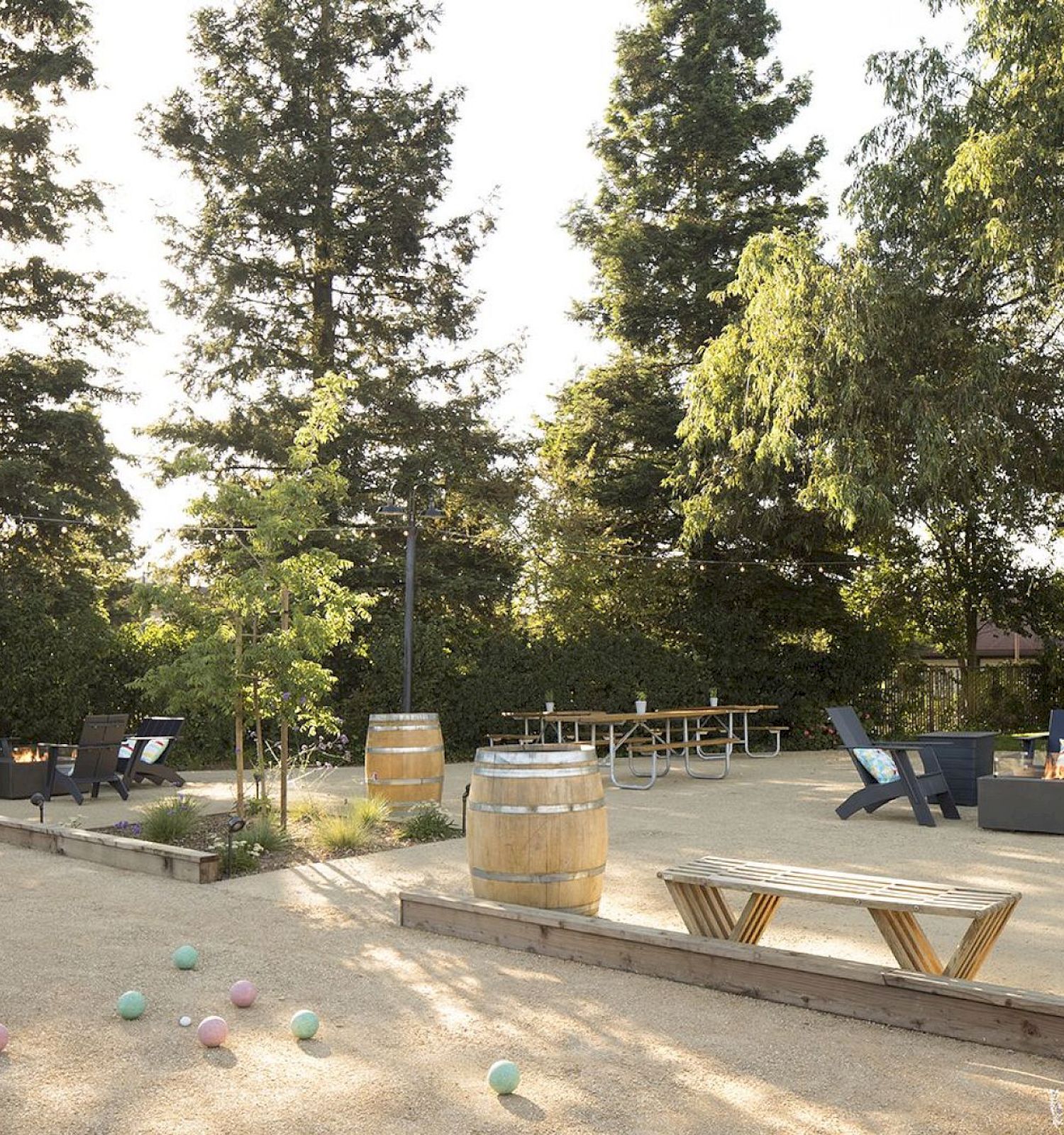 An outdoor space with seating areas, tables, chairs, barrels, and colorful balls on the ground, surrounded by tall trees and greenery.