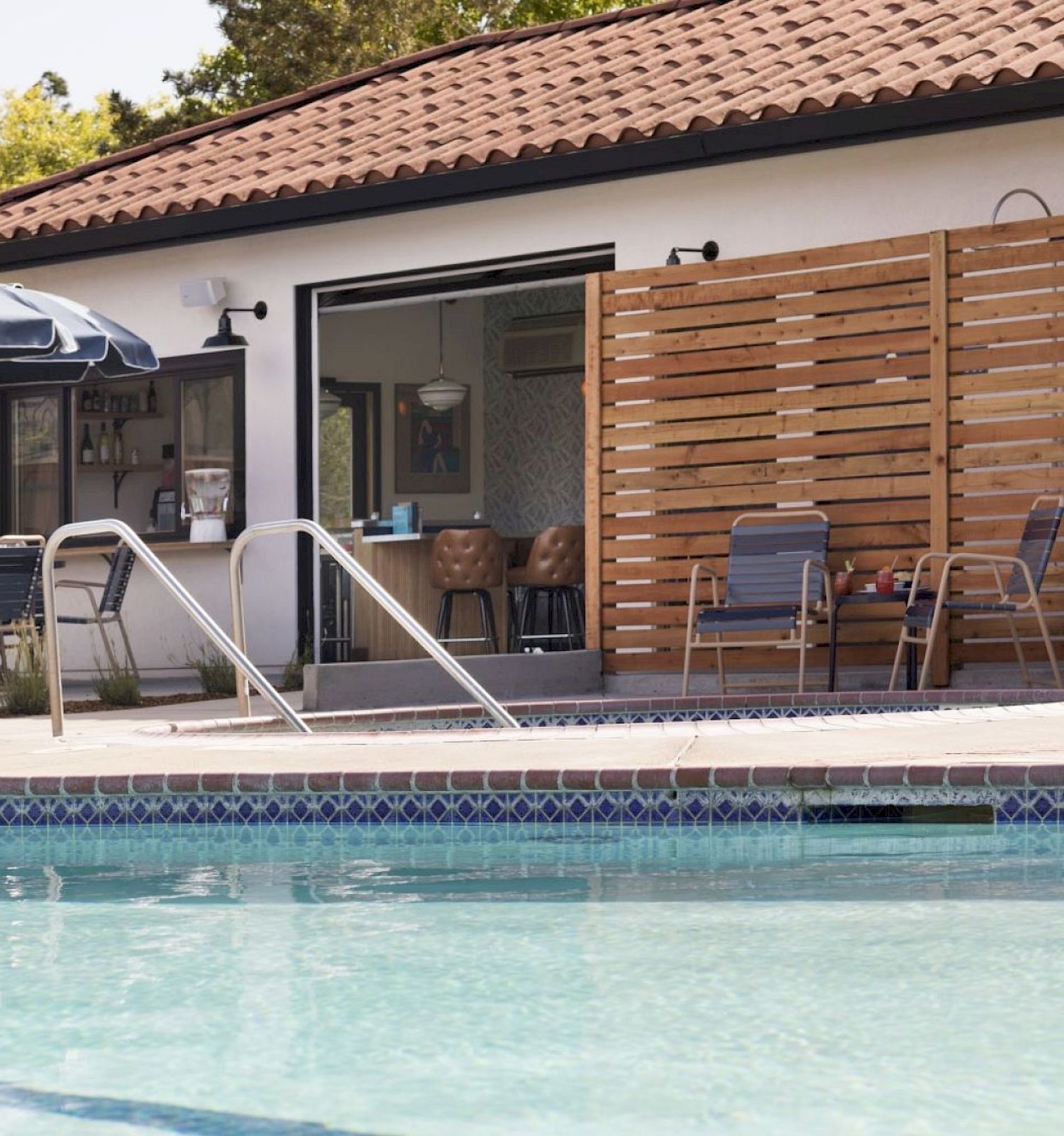 A poolside area with blue umbrellas, lounge chairs, and a building with a red-tiled roof in the background.