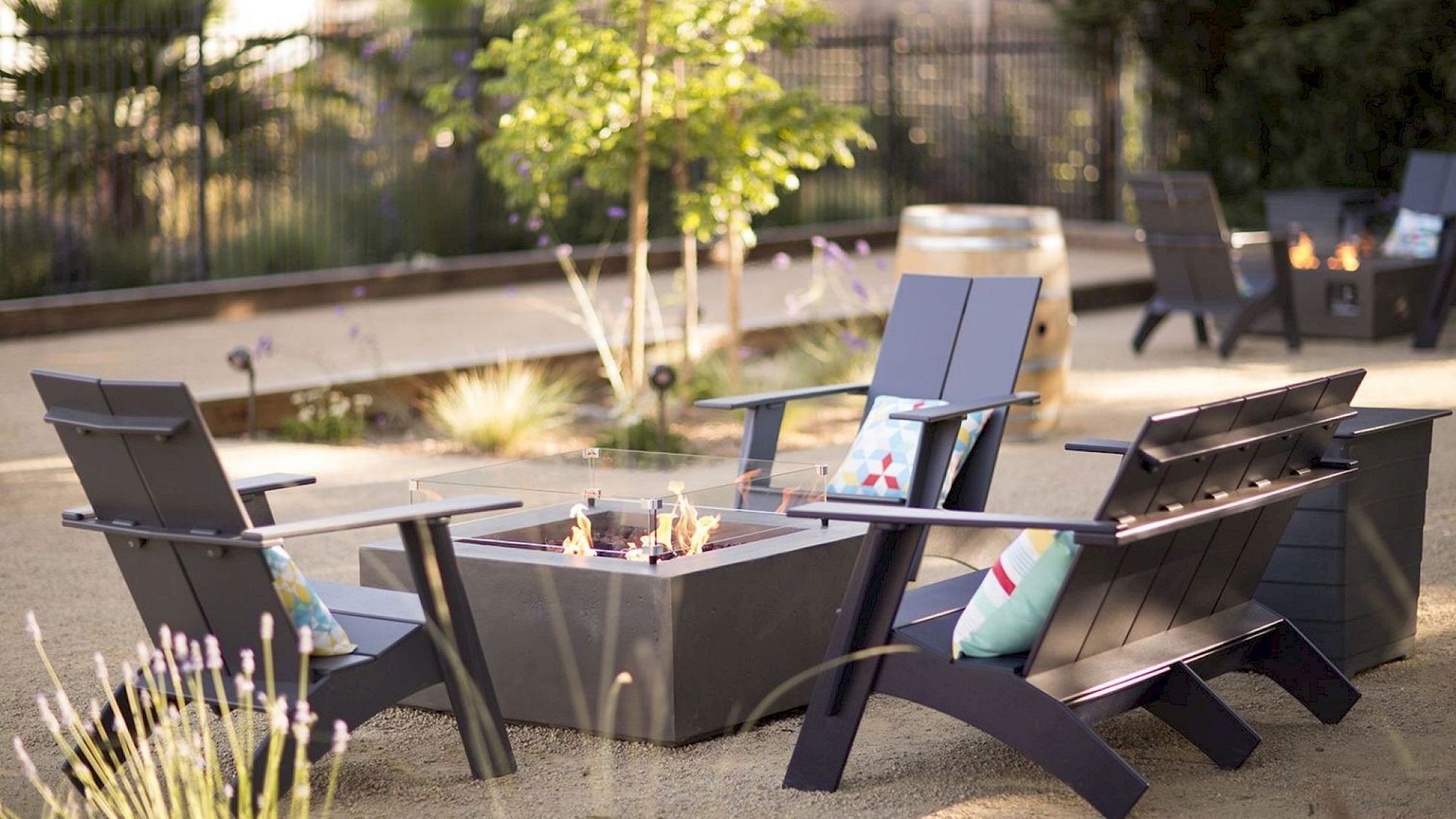 Outdoor seating area with dark chairs, a fire pit, pillows, and ambient lighting, surrounded by plants and trees.