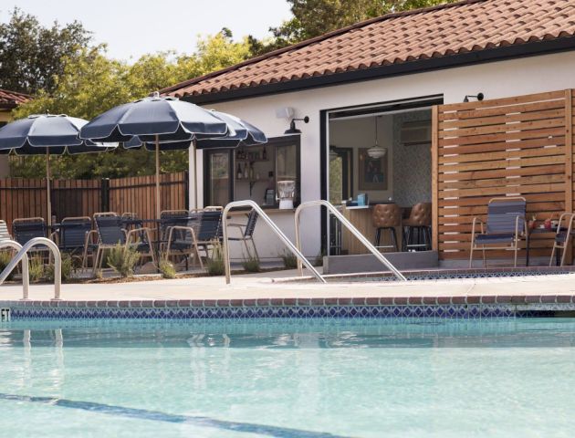 A swimming pool with clear water, lounge chairs, blue umbrellas, and a building with a tiled roof and outdoor seating area in the background.