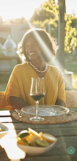 Three people are sitting at an outdoor table, enjoying wine and conversation, with sunlight filtering through trees, giving a warm ambiance.