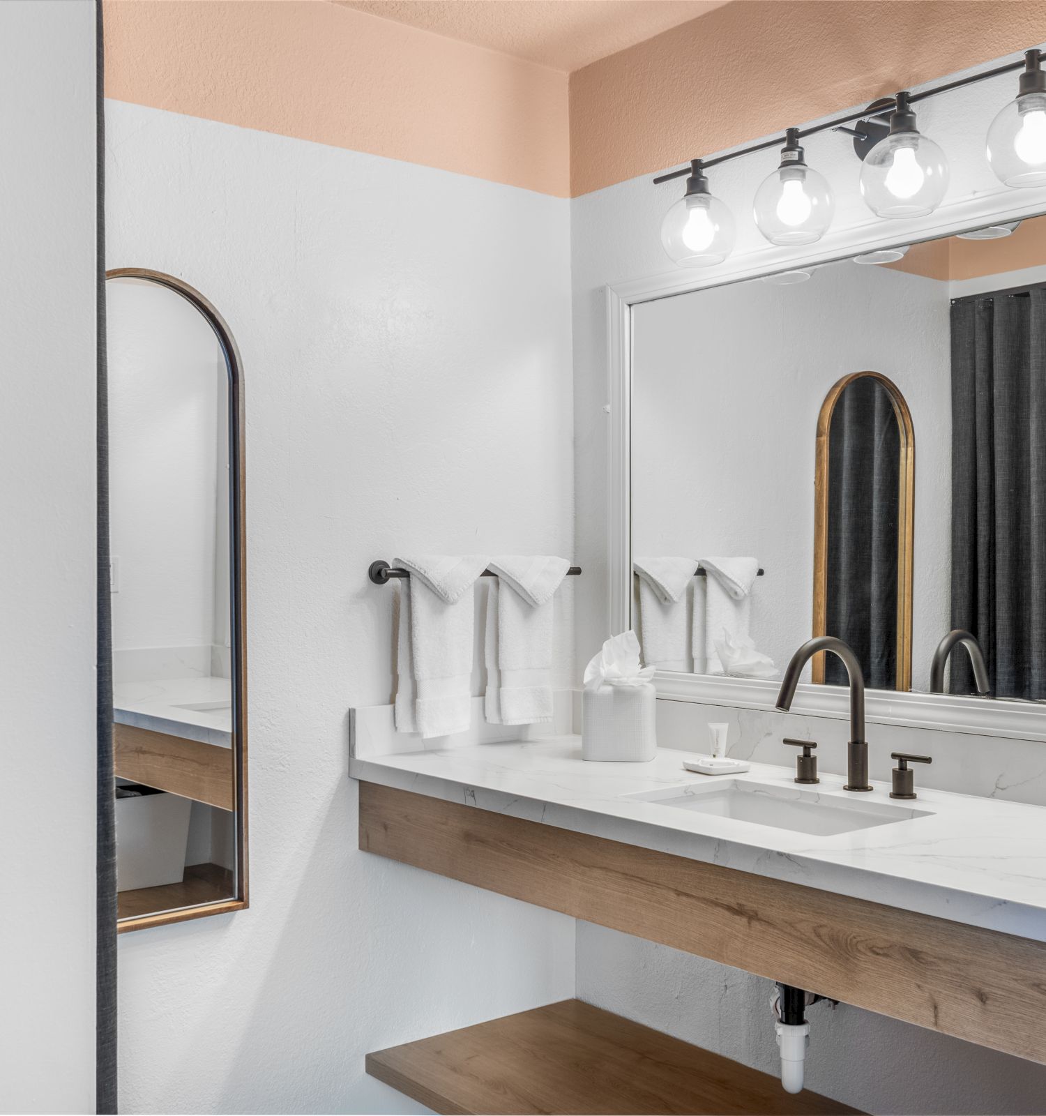 The image shows a modern bathroom with a large mirror, a countertop with a sink, three light bulbs above, and towels hanging on the wall.