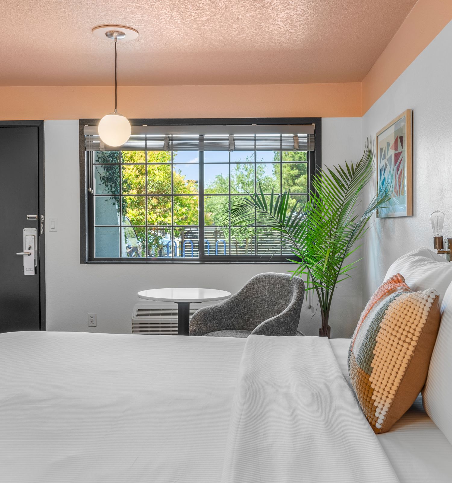 A modern hotel room with a large bed, stylish decor, a window view, potted plant, and a small seating area with a table and chair.