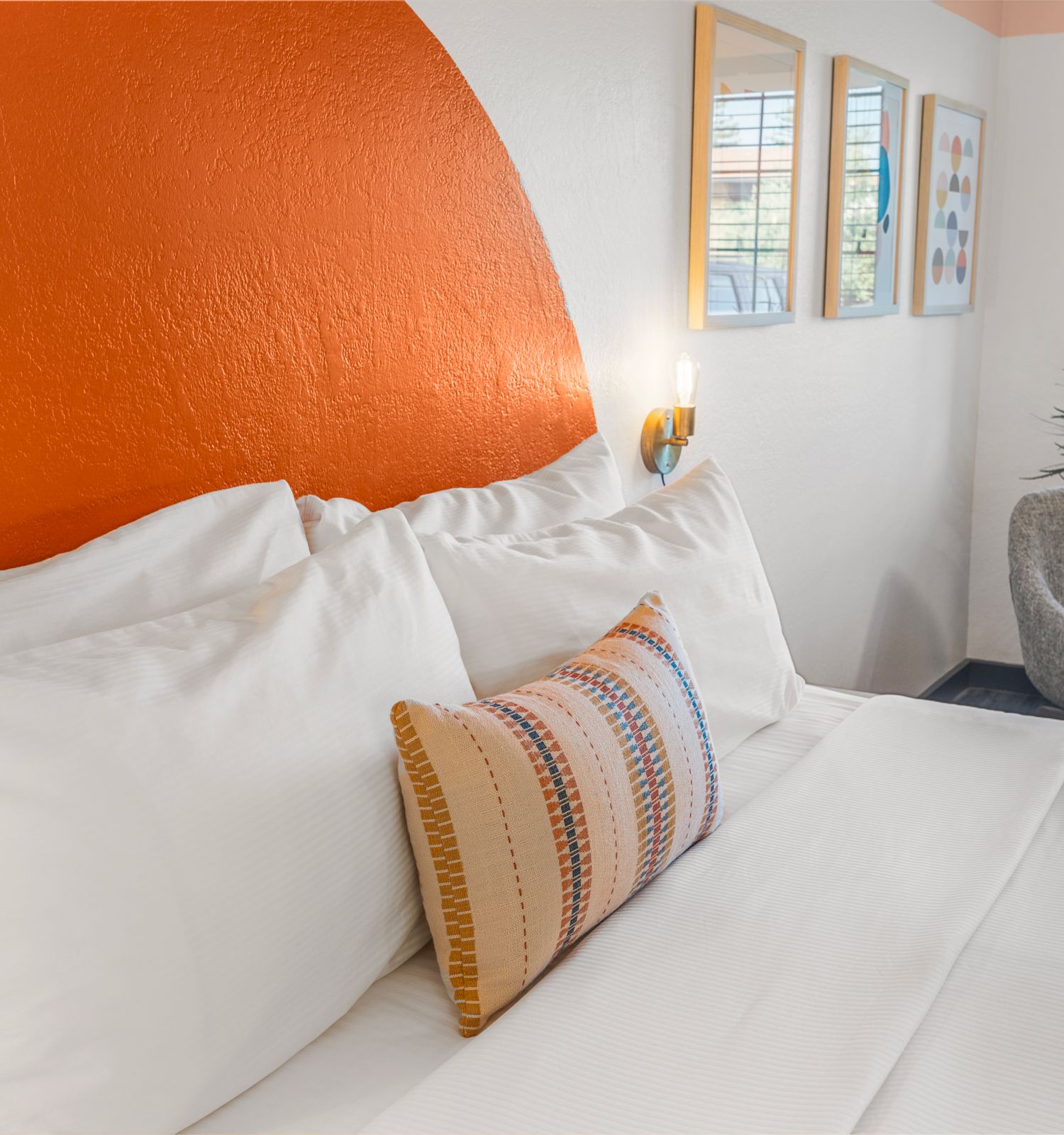 A neatly made bed with white linens and a decorative pillow is against an orange accent wall in a bright room with a chair and window.