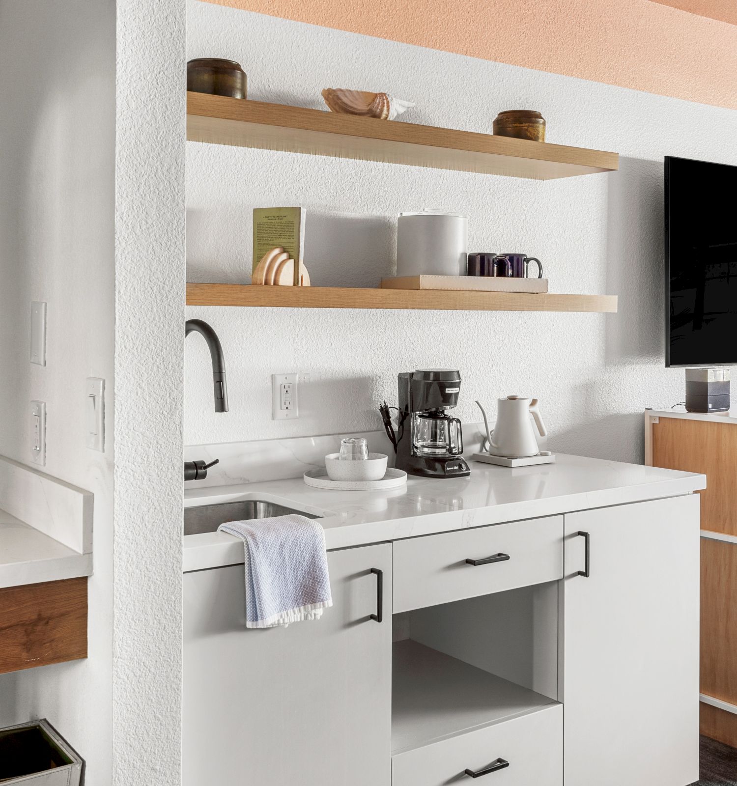 A modern kitchenette setup with a sink, open shelves, coffee maker, cups, a TV, and a mini-fridge in a bright room with a minimalist design.