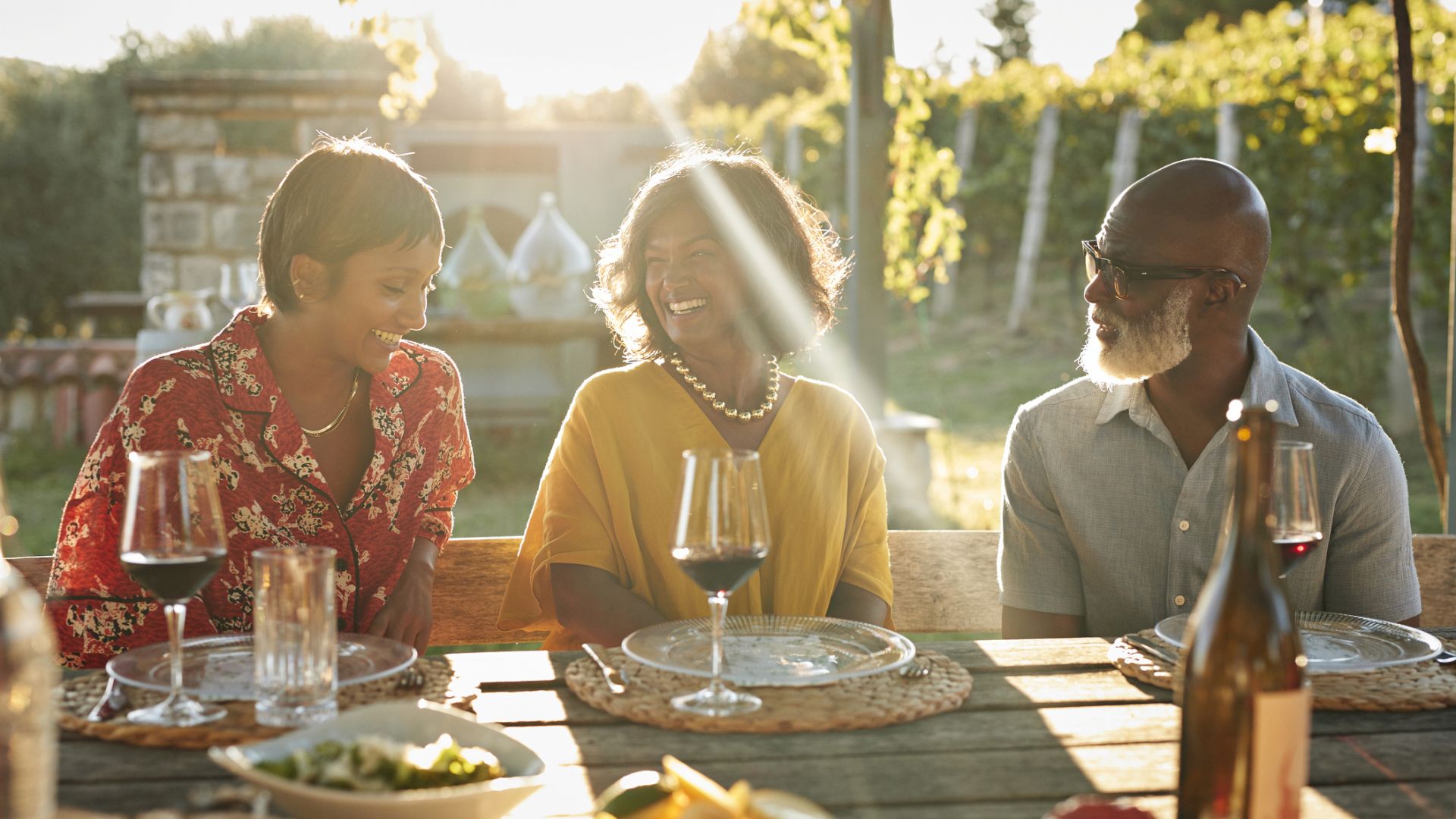 Three people are sitting outdoors at a table, enjoying a meal with wine and looking happy as they share a conversation in the sunlit setting.